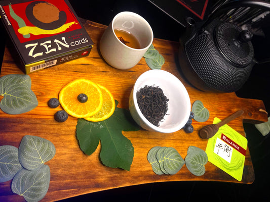 Tea-making setup with Georgian Black Loose Leaf Tea in a white cup, a box of 'Zen' cards, and a cast iron teapot on a wood block surface.