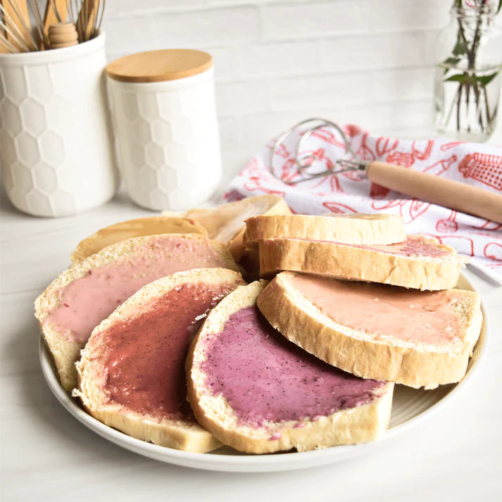 Slices of bread with several different flavored honey on slices of bread on an ecru colored plate, with wood kitchen utensils in the background.