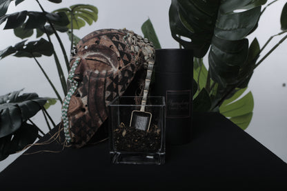 Black loose leaf tea blend in a glass container next to an African mask, on a black table with a  large leaf plant in the background.  