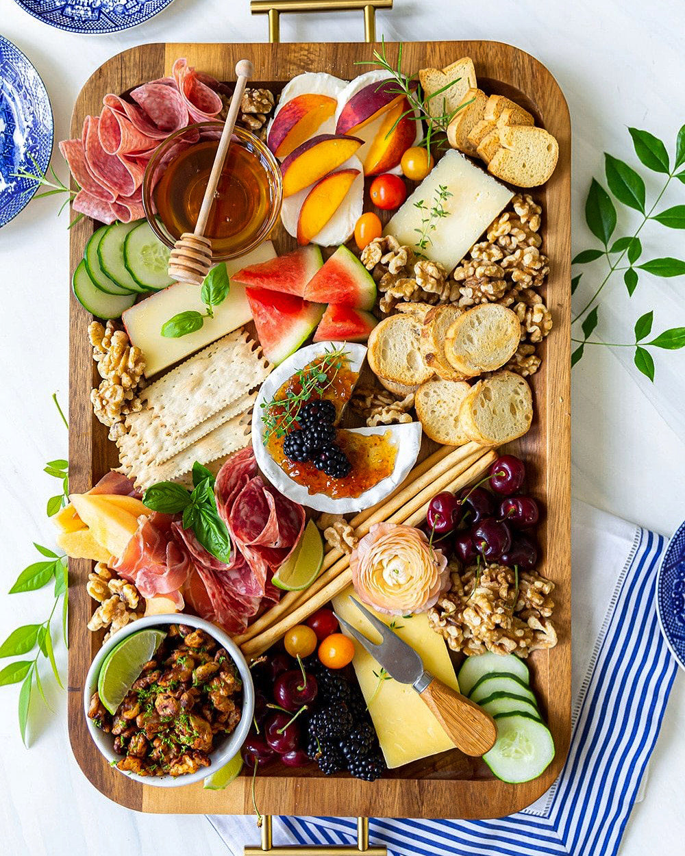 Wooden charcuterie board with various foods including meats, cheeses, fruits, and bread on a white surface.