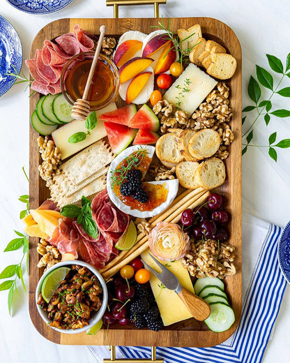 Wooden charcuterie board with various foods including meats, cheeses, fruits, and bread on a white surface.