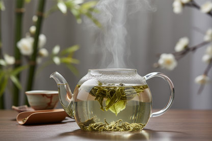 Jasmine Reverie flower unfurling in a glass tea pot. Celedene green color. In the background, ceramic tea cup, scooper, bamboo and delicate white flowers.