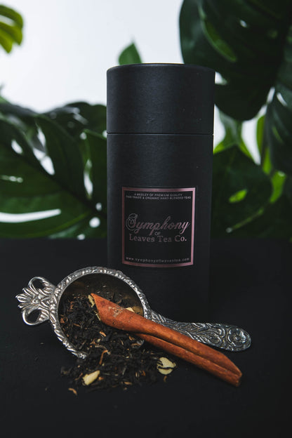 Black cylindrical container with rose gold label on a dark surface with black lapsang souchong tea leaves and cinnamon sticks.