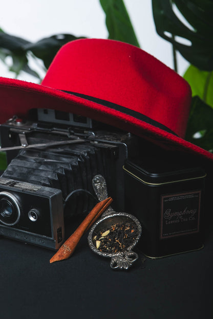 Red hat, vintage camera, black tin with brand name, and decorative item on a dark surface with green leaves in the background.