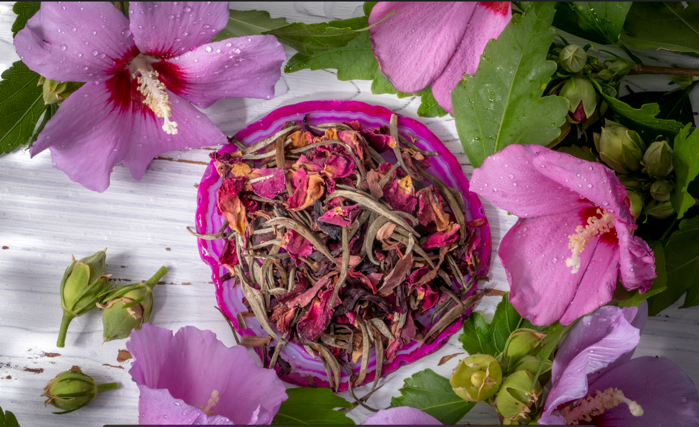 Bai Long Xu long white tea leaves, blended with dried roses, lavender and jasmine. The blend is in a pink container surrounded by fresh pink flowers and green leaves on a white wooden surface.