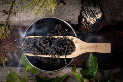 Wooden scoop filled with smoked oolong tea leaves on a rustic wooden surface with greenery.