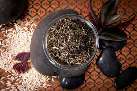 Thai loose oolong tea leaves in a black pot on a patterned surface with stones, rice grains and leaves.