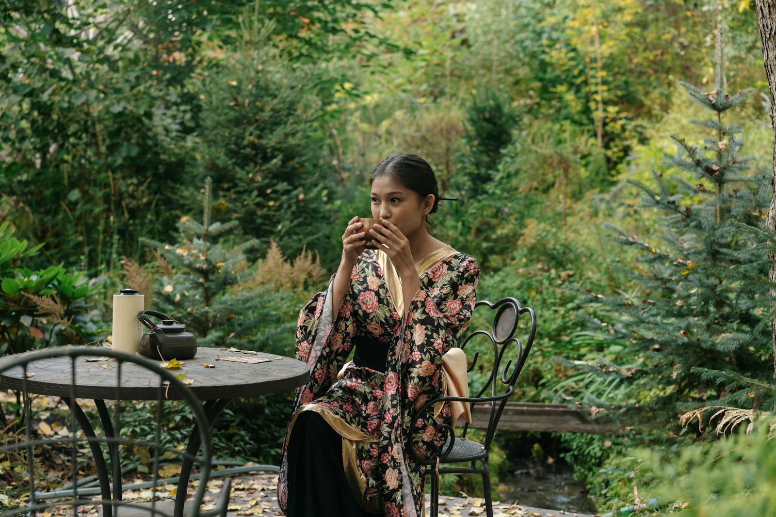 Asian woman wearing a floral kimono, sipping tea in a tranquil garden setting.